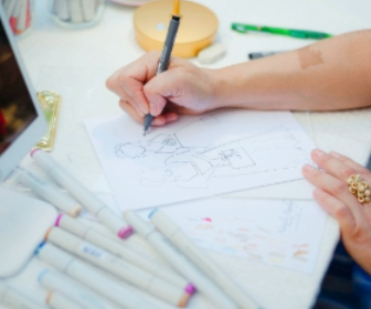 Close-up of artist Natalie Russo drawing a custom guest portrait at a live event, with her hand sketching in pen and ink on a white paper. The table is filled with colorful markers, showcasing the creative process behind her live event art.