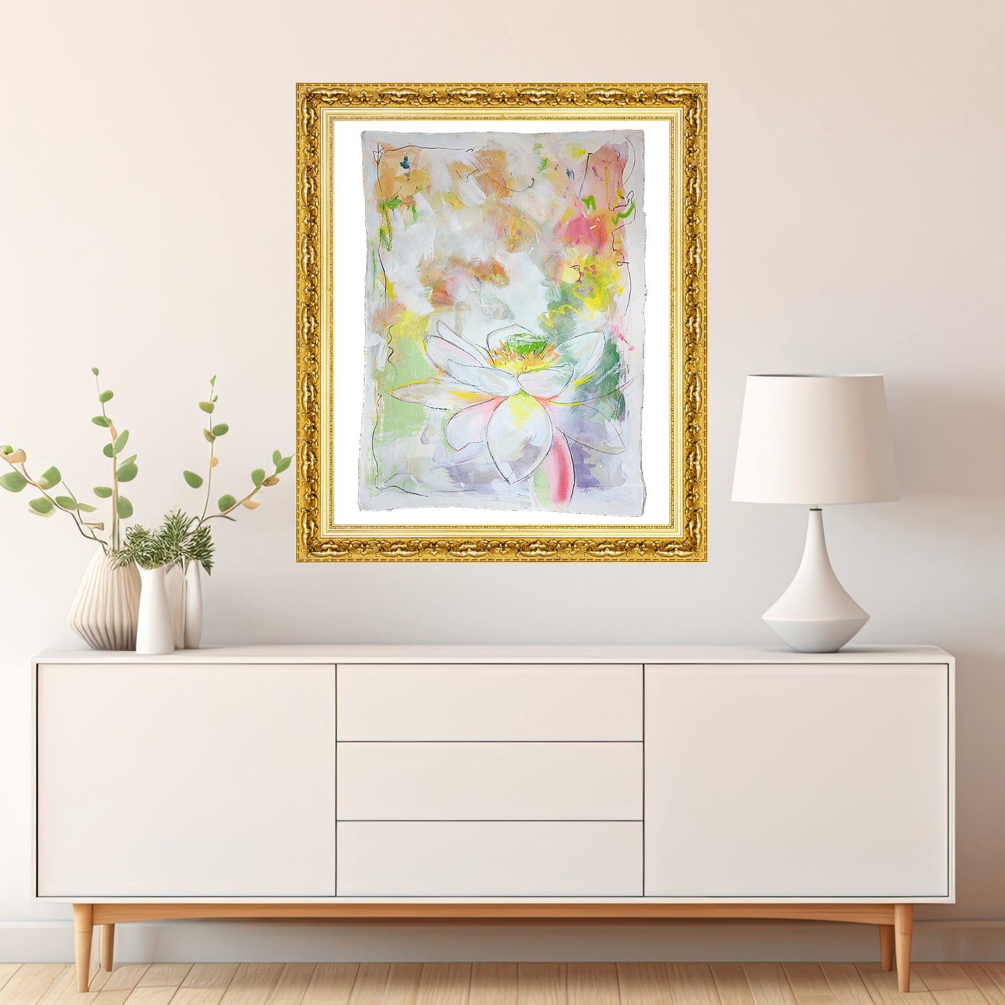 Original mixed media painting titled 'Serene Lotus Blossom' by Natalie Russo, displayed in a gold frame above a modern white sideboard with a lamp and decorative vase. The artwork features a delicate lotus flower in soft pastel tones with a touch of glitter, adding a serene and elegant touch to the interior.